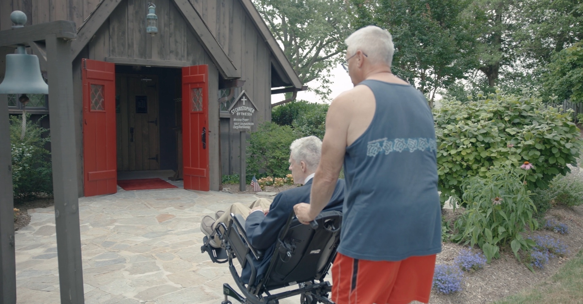 A caregiver pushing an older man in a wheelchair outdoors on a paved path near a golf course